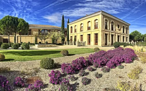 Finca El Caño, un edificio amarillo con arcos y balcones, rodeado de jardines verdes, arbustos y flores moradas bajo un cielo azul con nubes.