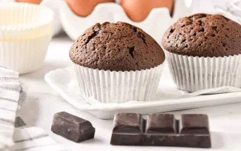 Dos magdalenas de chocolate recién horneadas, listas para disfrutar. Huevos y papelillos de repostería al fondo.