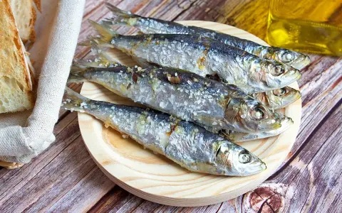 Cinco sardinas al horno, saladas y doradas, reposan en un plato de madera junto a pan rústico y una botella de aceite.