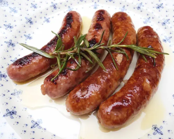 Salchichas al vino Monsieur Cuisine, doradas y jugosas, con romero fresco sobre un plato blanco con motivos azules.