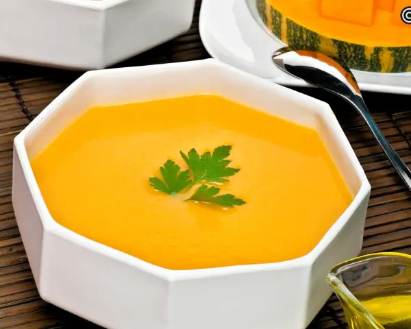 Un cremoso y vibrante **pure de calabaza** en un cuenco blanco, decorado con perejil fresco. Al fondo, un trozo de calabaza y una cuchara.