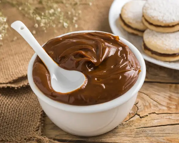 Un tazón blanco lleno de cremoso dulce de leche, con una cuchara blanca sumergida. Al fondo, alfajores espolvoreados con azúcar glas.