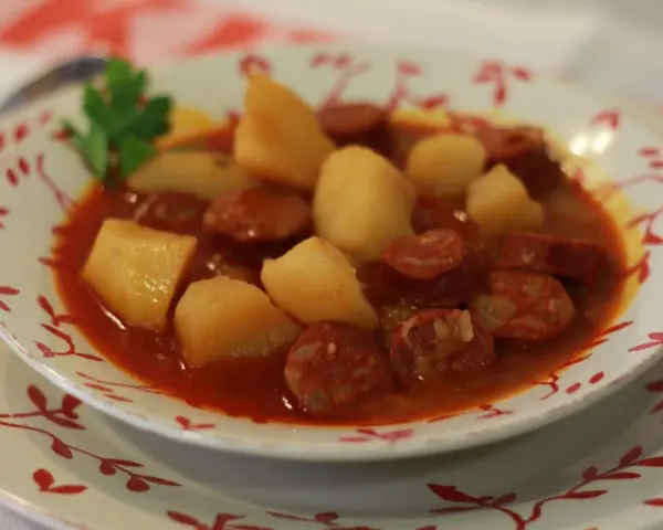 Un plato humeante de patatas a la riojana, con trozos de chorizo y patata en una salsa rojiza, adornado con una ramita de perejil.