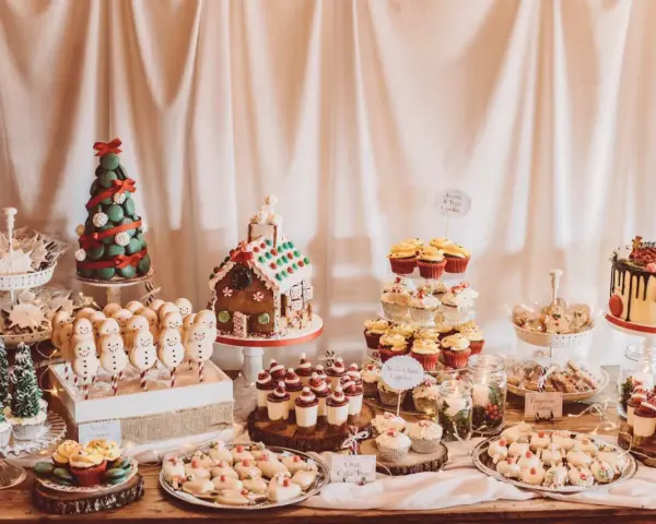 Mesa llena de postres navideños: casa de jengibre, árbol de macarons, muñecos de nieve, cupcakes y galletas.