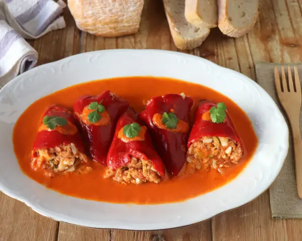 Cinco pimientos rellenos, cubiertos con salsa y perejil, servidos en un plato blanco con pan y cubiertos de madera.