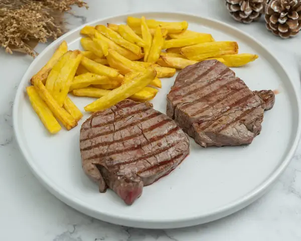 Dos jugosos solomillos de ternera a la plancha, con marcas de parrilla, acompañados de patatas fritas doradas.
