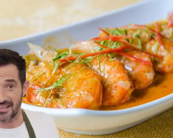 Un hombre sonriente presenta un plato de gambas al ajillo, con gambas jugosas en una salsa naranja, adornadas con finas tiras de chile rojo y hierbas verdes.