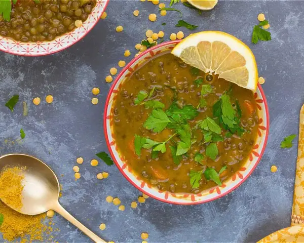 Un tazón de lentejas al curry, adornado con perejil fresco y una rodaja de limón. Una cuchara dorada con cúrcuma completa la escena.
