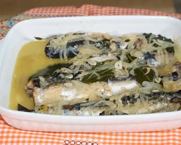 Caballa en escabeche servida en una fuente blanca, con cebolla, laurel y un líquido dorado.