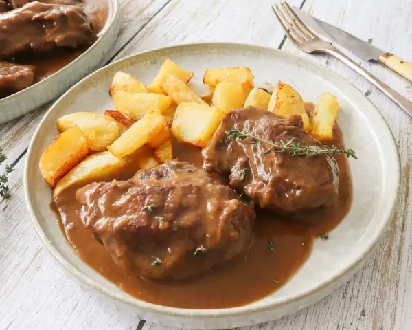 Dos tiernas porciones de pata de ternera guisada en su jugo, acompañadas de patatas rústicas fritas y una ramita de tomillo.