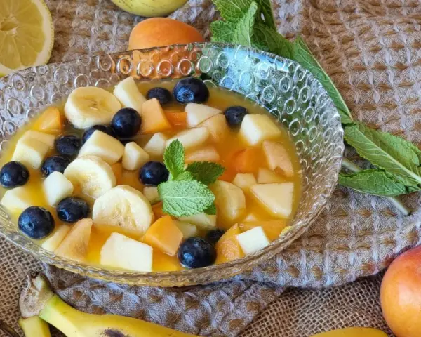 Deliciosa macedonia de frutas con plátano, arándanos, melocotón y manzana en un cuenco de cristal, decorada con menta.
