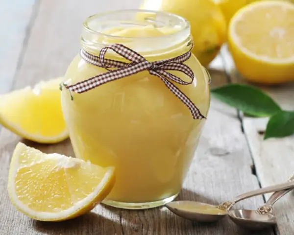 Tarro de lemon curd casero, adornado con un lazo a cuadros, junto a limones frescos y hojas verdes sobre una mesa de madera.