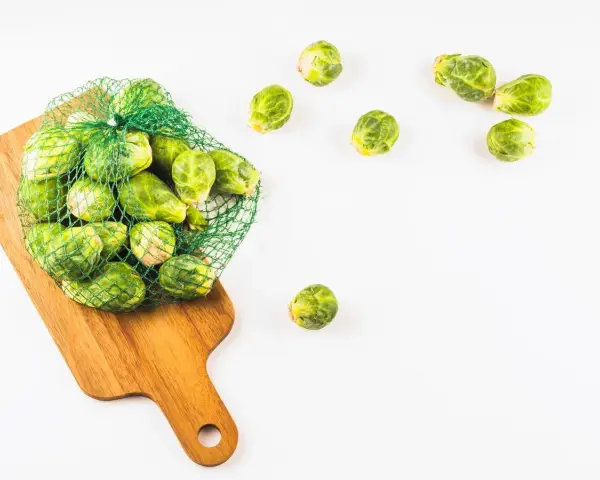 Una bolsa de malla verde llena de coles de Bruselas frescas descansa sobre una tabla de cortar de madera. Algunas coles de Bruselas sueltas se esparcen alrededor.