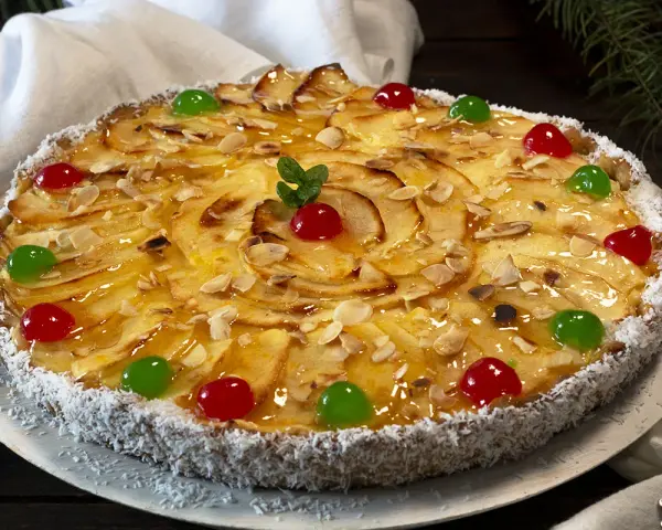 Deliciosa tarta de manzana cubierta de almendras laminadas y cerezas rojas y verdes. El borde está cubierto de coco rallado.