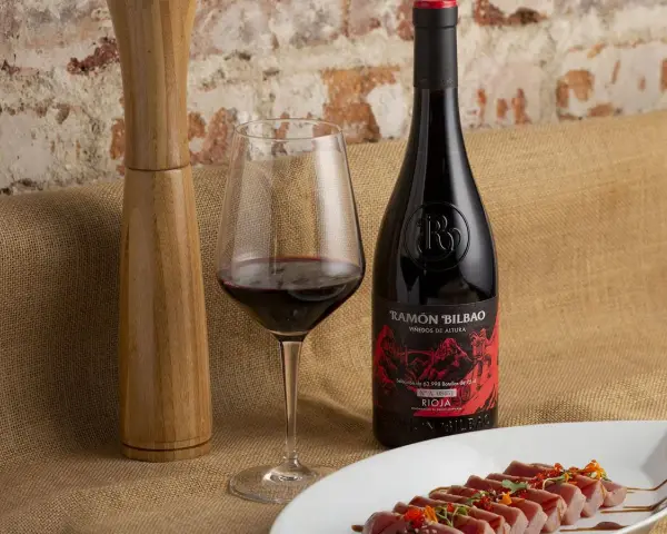 Una copa de vino seco, una botella de Ramón Bilbao Rioja y un plato de atún sellado, listos para una cena.