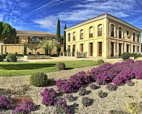 Finca El Caño, un edificio amarillo con arcos y balcones, rodeado de jardines verdes, arbustos y flores moradas bajo un cielo azul con nubes.