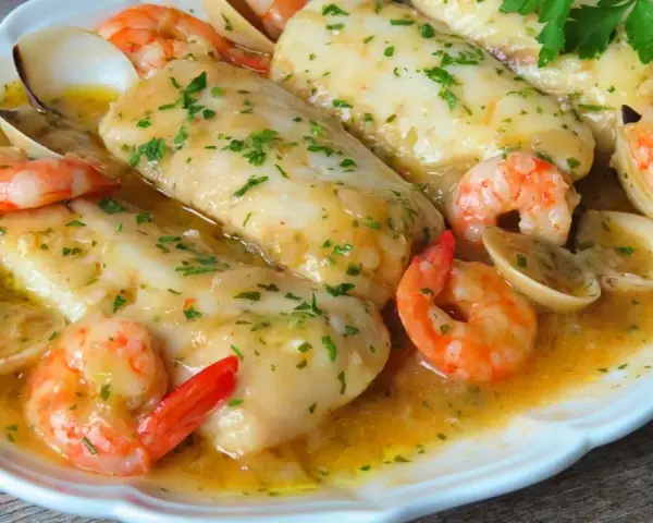 Merluza a la marinera con gambas y almejas, todo bañado en una deliciosa salsa dorada y espolvoreado con perejil fresco.
