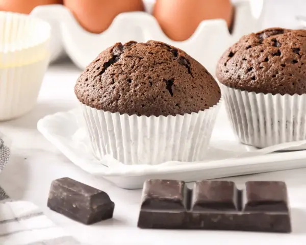 Dos magdalenas de chocolate recién horneadas, listas para disfrutar. Huevos y papelillos de repostería al fondo.