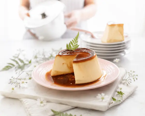 Delicioso flan de queso con caramelo líquido, decorado con una ramita verde. Se apilan más porciones en el fondo.