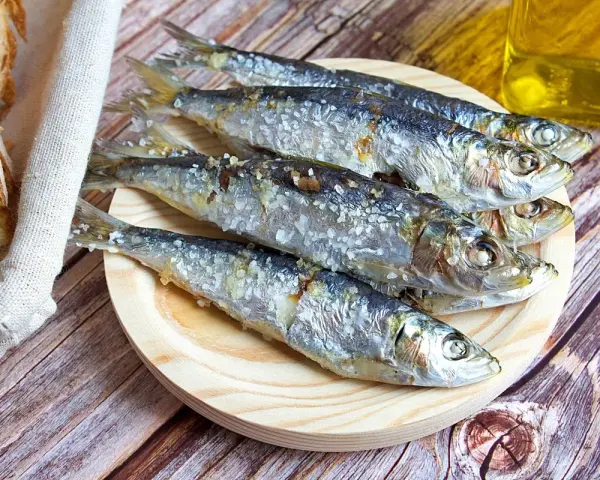 Cinco sardinas al horno, saladas y doradas, reposan en un plato de madera junto a pan rústico y una botella de aceite.