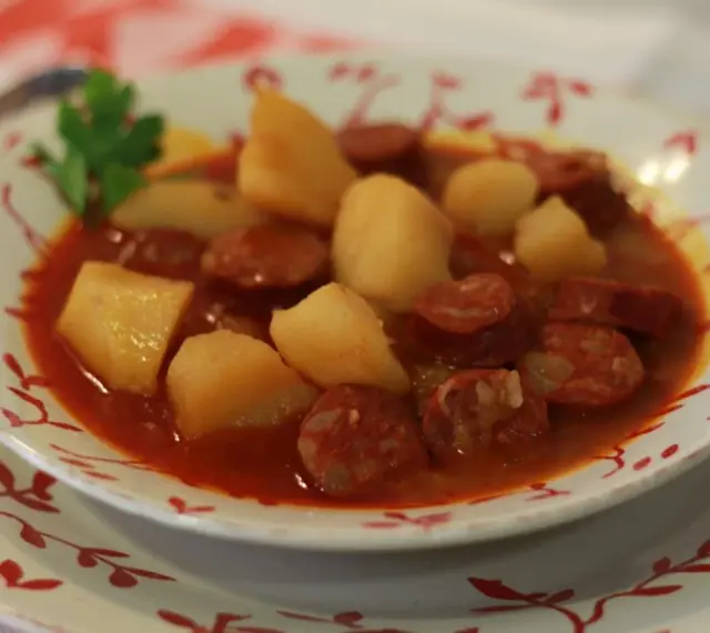 Un plato humeante de patatas a la riojana, con trozos de chorizo y patata en una salsa rojiza, adornado con una ramita de perejil.
