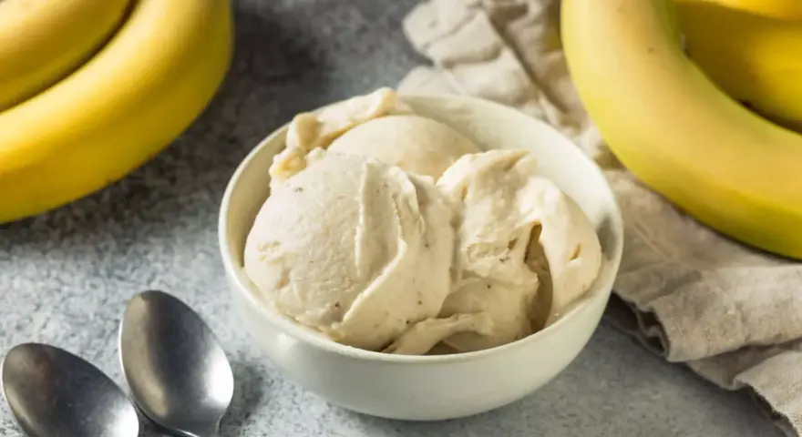 Un cremoso helado de plátano en un tazón blanco, con plátanos amarillos frescos al lado. ¡Perfecto para un postre refrescante!