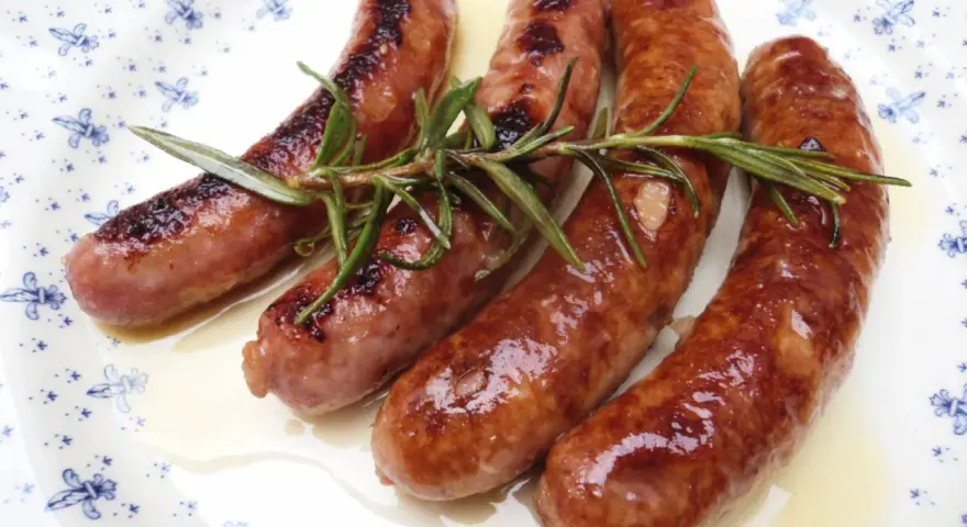 Salchichas al vino Monsieur Cuisine, doradas y jugosas, con romero fresco sobre un plato blanco con motivos azules.