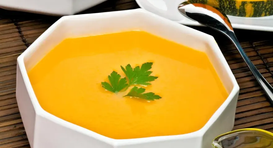 Un cremoso y vibrante **pure de calabaza** en un cuenco blanco, decorado con perejil fresco. Al fondo, un trozo de calabaza y una cuchara.