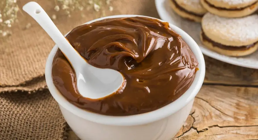 Un tazón blanco lleno de cremoso dulce de leche, con una cuchara blanca sumergida. Al fondo, alfajores espolvoreados con azúcar glas.