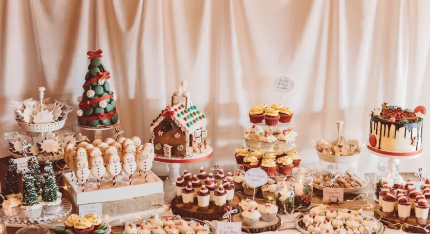 Mesa llena de postres navideños: casa de jengibre, árbol de macarons, muñecos de nieve, cupcakes y galletas.