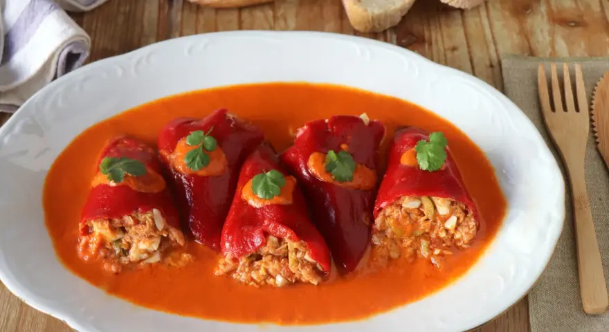 Cinco pimientos rellenos, cubiertos con salsa y perejil, servidos en un plato blanco con pan y cubiertos de madera.
