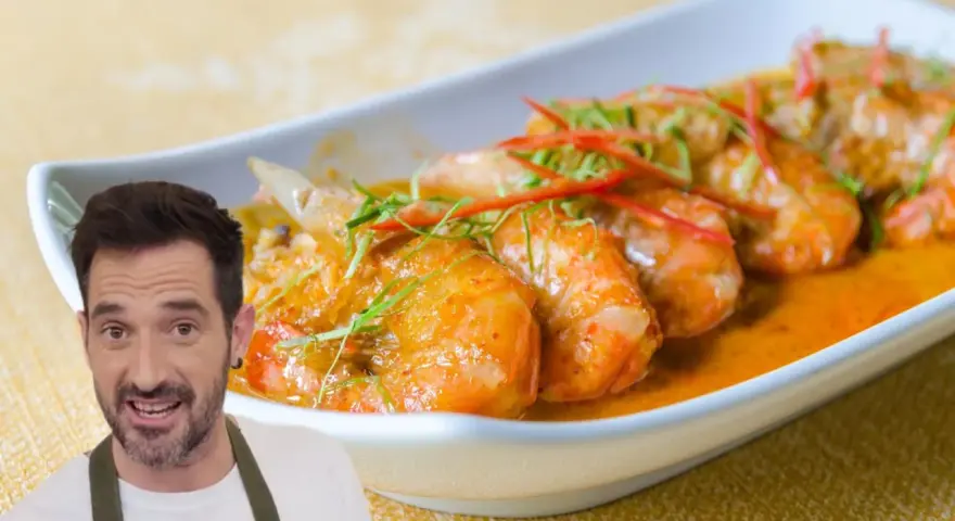 Un hombre sonriente presenta un plato de gambas al ajillo, con gambas jugosas en una salsa naranja, adornadas con finas tiras de chile rojo y hierbas verdes.
