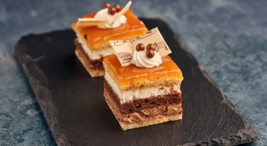 Deliciosa tarta San Marcos en porciones, con capas de bizcocho, crema y cobertura anaranjada, decorada con nata y notas musicales de chocolate.