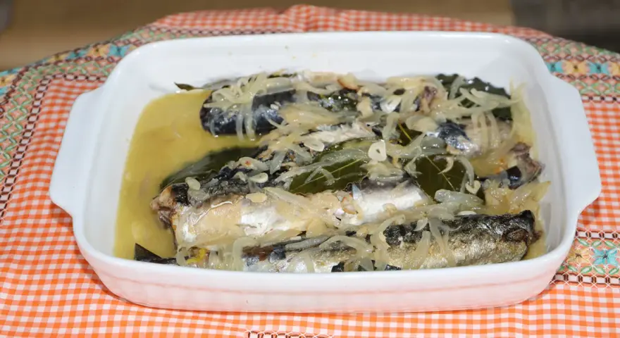 Caballa en escabeche servida en una fuente blanca, con cebolla, laurel y un líquido dorado.