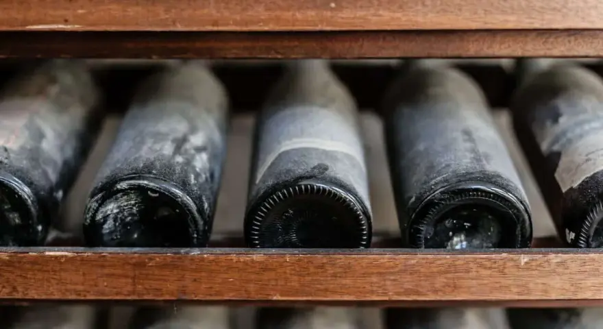Botellas de vino añejo en estantería de madera, cubiertas de polvo y con el sedimento del vino visible.