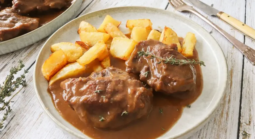 Dos tiernas porciones de pata de ternera guisada en su jugo, acompañadas de patatas rústicas fritas y una ramita de tomillo.