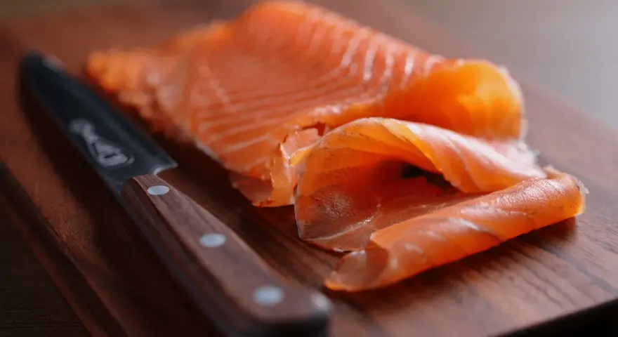 Finísimas lonchas de salmón marinado, listas para disfrutar, junto a un cuchillo de chef sobre tabla de madera.