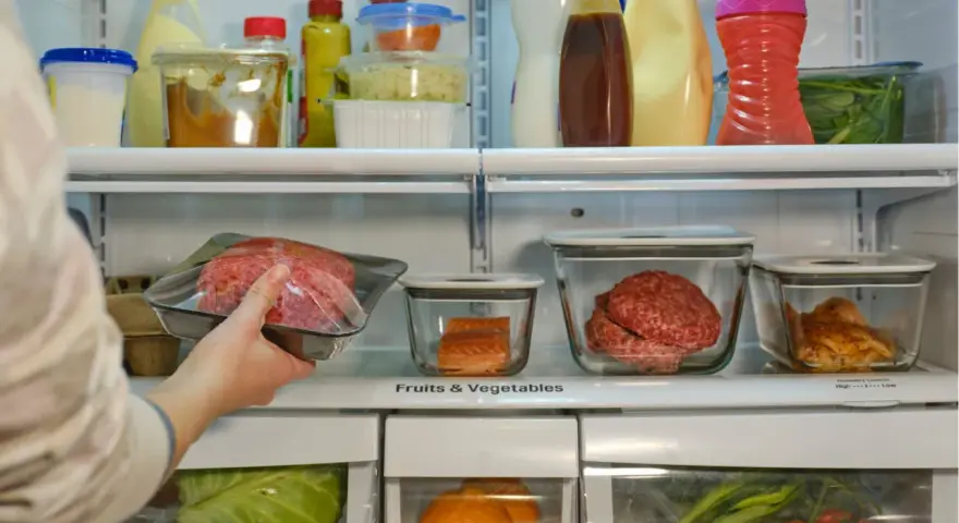 Mano colocando carne picada en la nevera. ¿Cuánto dura la carne en la nevera? También hay salmón, pollo y verduras.