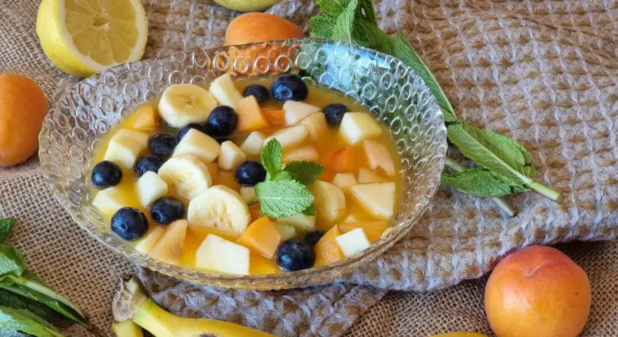 Deliciosa macedonia de frutas con plátano, arándanos, melocotón y manzana en un cuenco de cristal, decorada con menta.