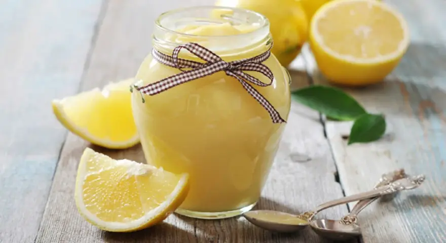 Tarro de lemon curd casero, adornado con un lazo a cuadros, junto a limones frescos y hojas verdes sobre una mesa de madera.