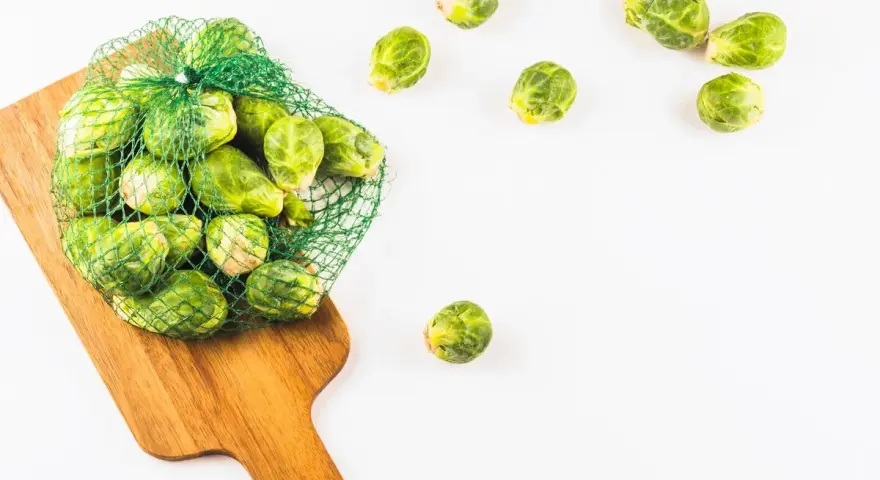 Una bolsa de malla verde llena de coles de Bruselas frescas descansa sobre una tabla de cortar de madera. Algunas coles de Bruselas sueltas se esparcen alrededor.