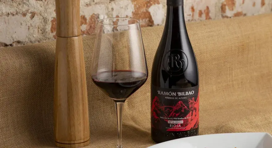 Una copa de vino seco, una botella de Ramón Bilbao Rioja y un plato de atún sellado, listos para una cena.