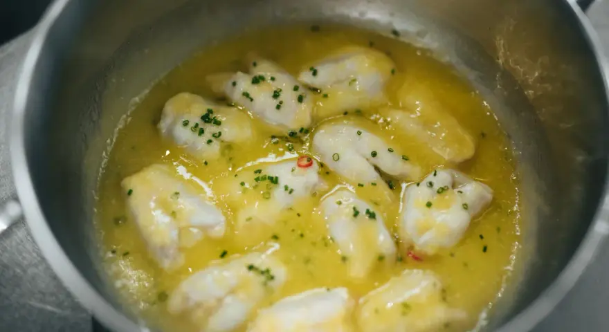 Trozo de bacalao al pil pil burbujeando en una sartén con aceite de oliva, ajo y perejil picado.
