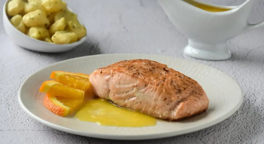 Filete de salmón a la naranja, servido con gajos de naranja y una salsa dorada. Acompañado de gnocchis en un cuenco.