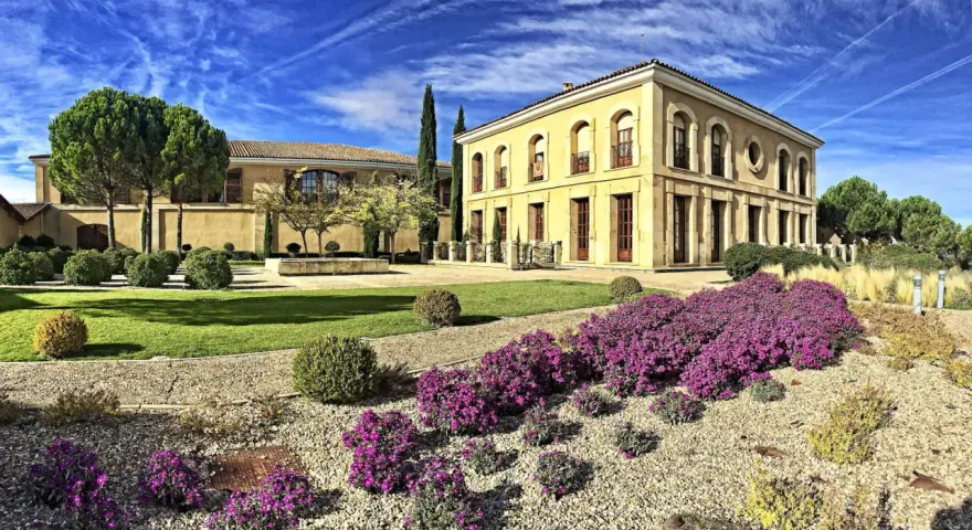 Finca El Caño, un edificio amarillo con arcos y balcones, rodeado de jardines verdes, arbustos y flores moradas bajo un cielo azul con nubes.