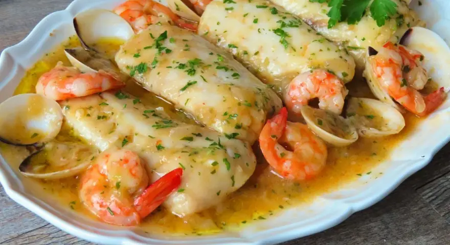 Merluza a la marinera con gambas y almejas, todo bañado en una deliciosa salsa dorada y espolvoreado con perejil fresco.