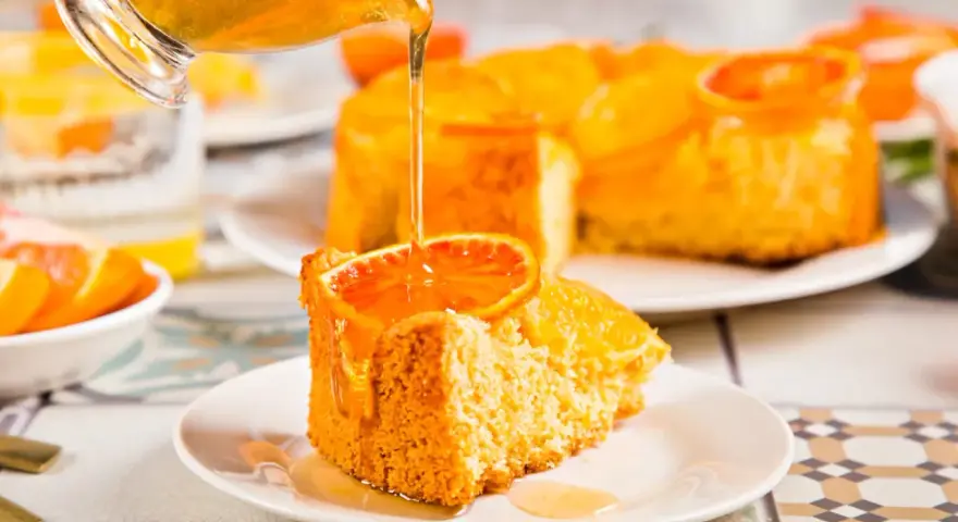 Un trozo de tarta de naranja esponjosa, decorado con una rodaja de naranja y bañado en un almíbar dorado.