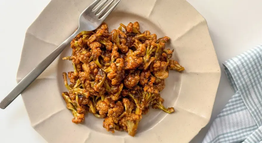 Coliflor glaseada, una de las mejores recetas con coliflor. Servida en plato beige con tenedor plateado y servilleta a cuadros.