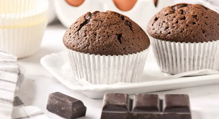 Dos magdalenas de chocolate recién horneadas, listas para disfrutar. Huevos y papelillos de repostería al fondo.