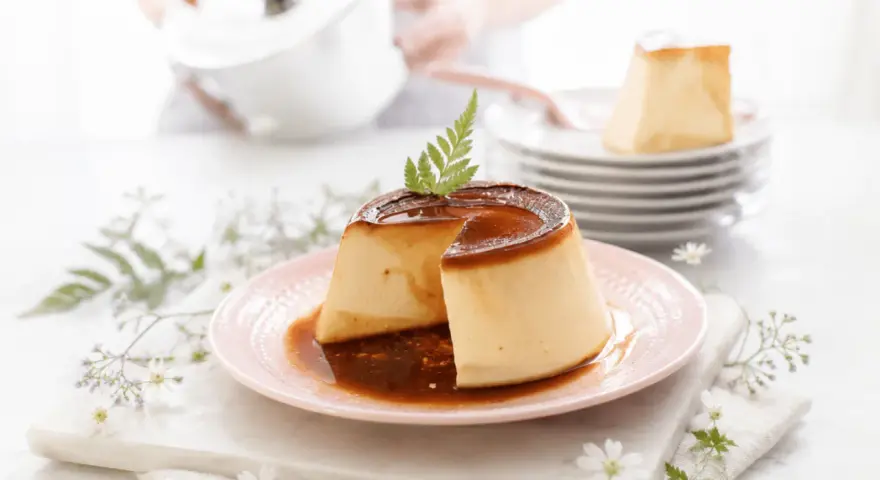 Delicioso flan de queso con caramelo líquido, decorado con una ramita verde. Se apilan más porciones en el fondo.